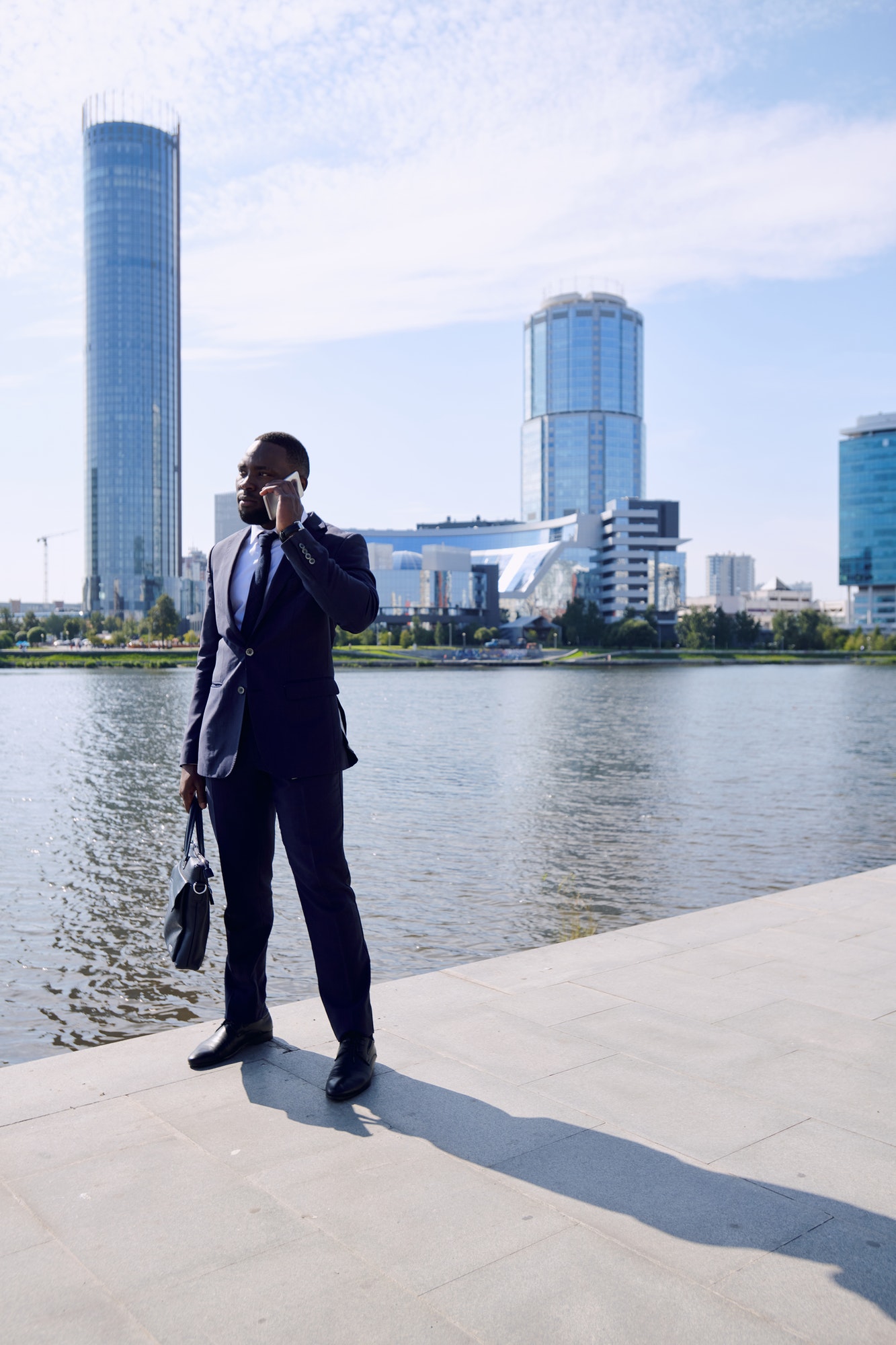 Young elegant African businessman or agent phoning against riverside and cityscape with skyscrapers